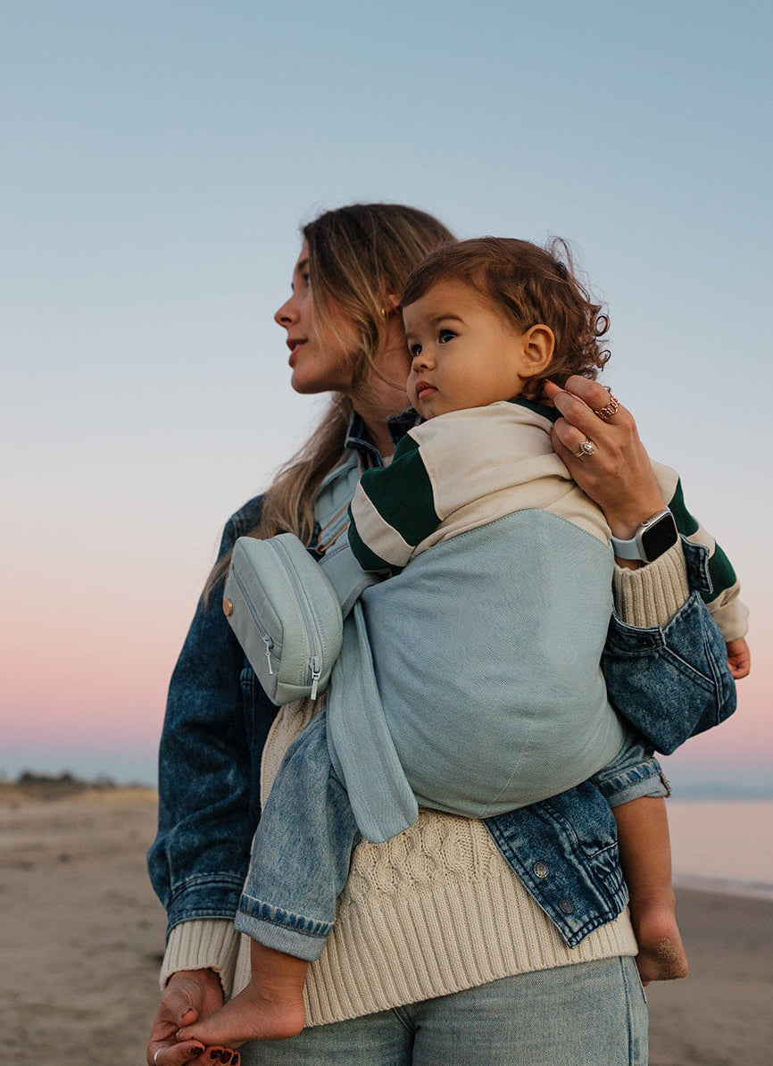 The Mabē Monarch Toddler Sling and matching Accessory Bag in Dream Wash Denim, which is solid light blue all-cotton fabric, worn by a woman and toddler.