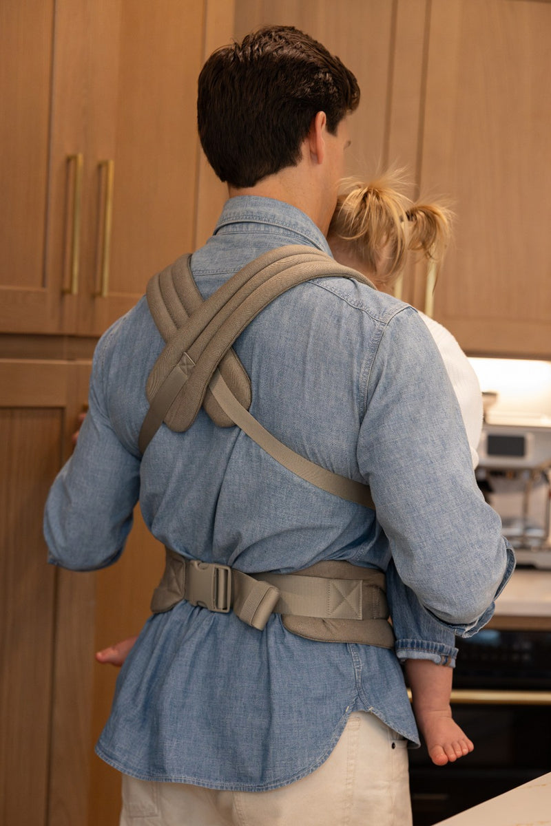 Man wearing the Mabē Monarch Buckle Carrier in Charles Herringbone, which is a delicate herringbone weave cotton/linen fabric in shades of brown. Back view.