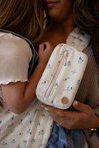 Woman wearing the Mabē Monarch Toddler Sling and Accessory Bag in Sunday Floral, which has solid white cotton/linen fabric with tiny sprigs of blue flowers, carrying an older toddler.