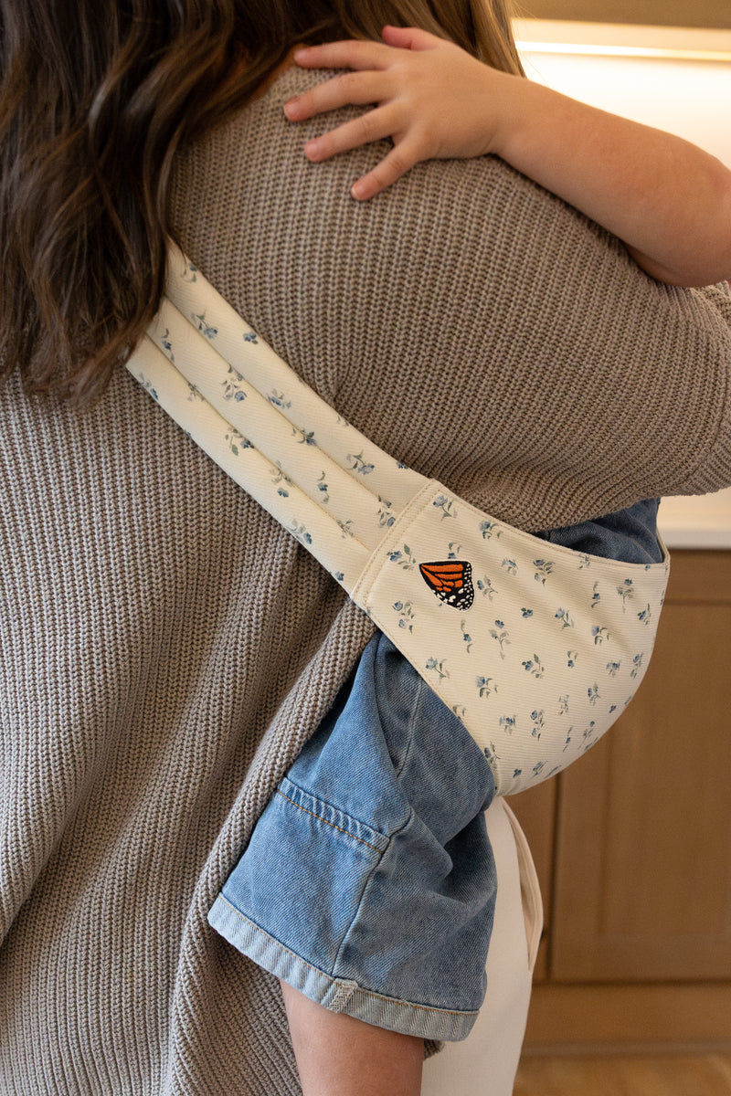 Unseen adult and toddler using a Mabē Monarch Toddler Sling in Sunday Floral, which is solid white cotton/linen with tiny sprigs of blue flowers.