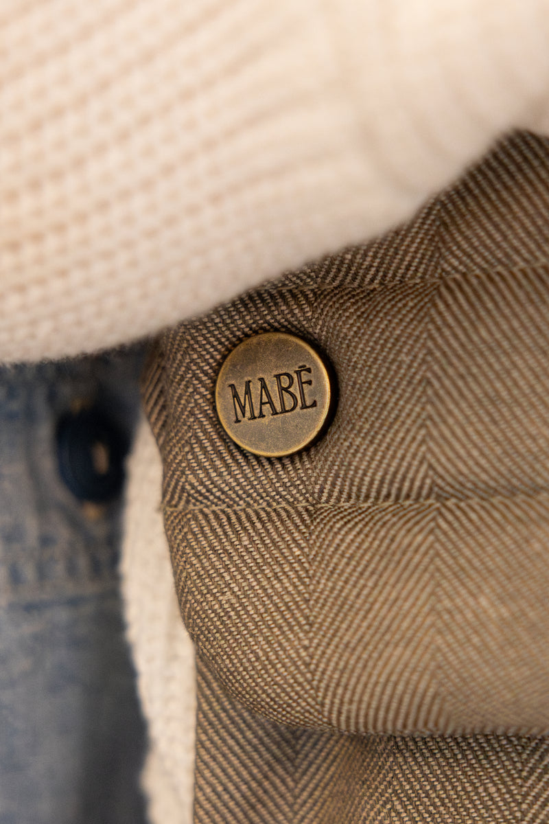 Close-up of the Mabē Monarch Buckle Carrier in Charles Herringbone with an antiqued bronze button labeled 'MABĒ.' The fabric is a brown herringbone weave cotton/linen.