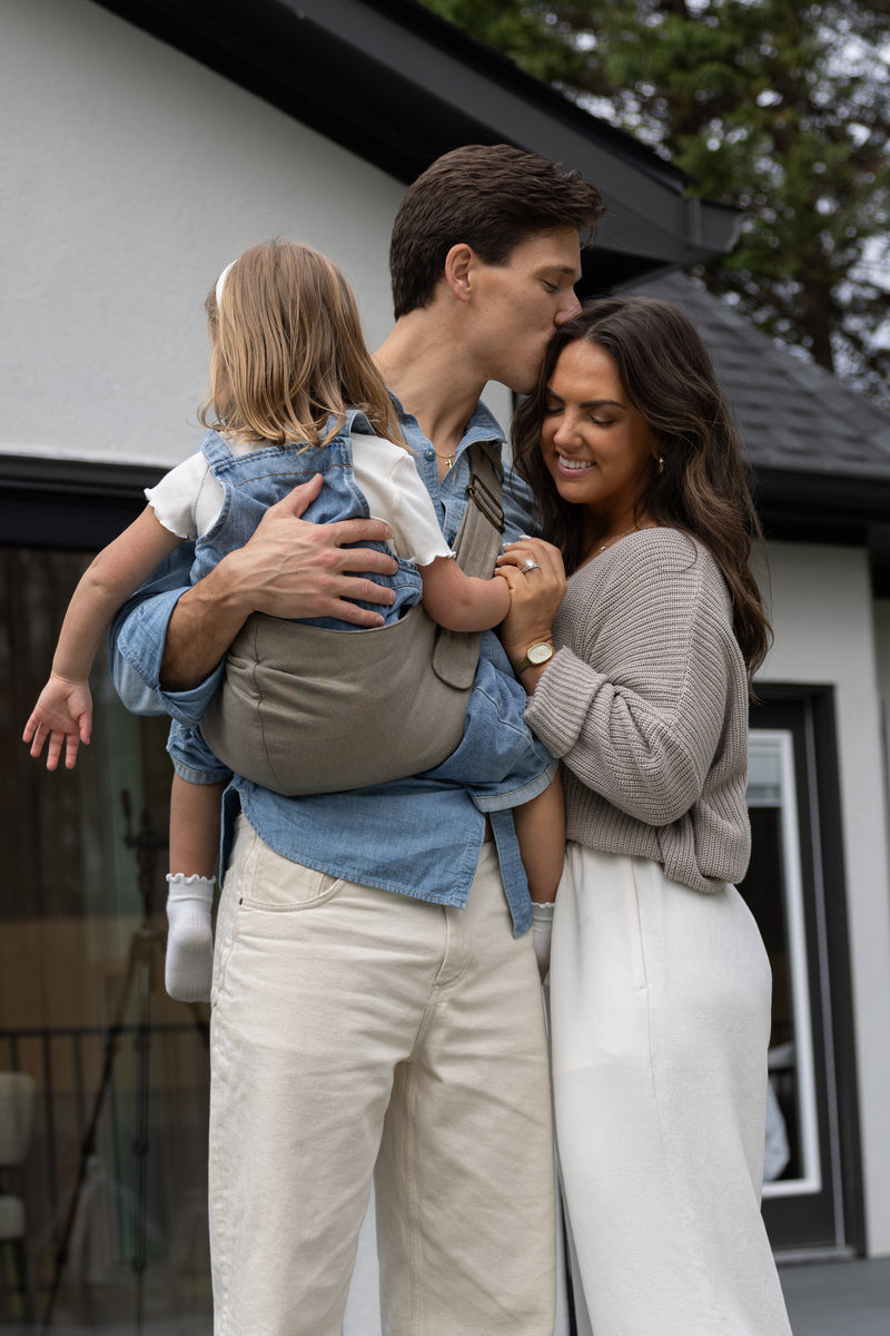 The Mabē Monarch Toddler Sling in Sunday Floral (solid white with tiny sprigs of blue flowers) and Mabē Monarch Buckle Carrier in Charles Herringbone (delicate herringbone pattern in shades of brown) worn by Liz and Zach Webber with their two children.