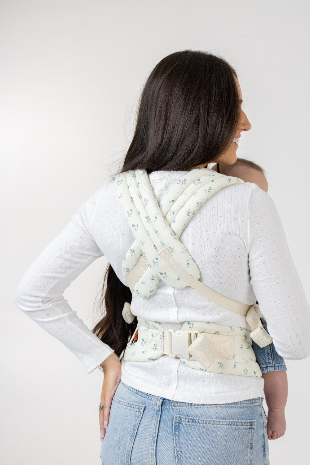 A woman with straight, dark hair carries a mostly unseen baby with Buckle Carrier in Sunday Floral, which is solid white with tiny sprigs of blue flowers. She is facing away from the viewer. The woman wears a fitted white long-sleeved shirt and light-wash jeans and is shown from her bottom up. The background is solid white. Only the crossed straps and waistband of the carrier are visible across her back.