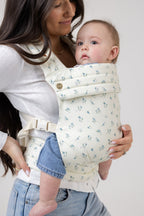A tanned-skinned woman with straight, dark brown hair carries a light-skinned baby with brown hair in the Mabē Monarch Buckle Carrier in Sunday Floral, which is solid white with tiny sprigs of blue flowers. The woman wears a fitted white long-sleeved shirt and light-wash jeans and is shown from just above the hips up. The background is solid white.