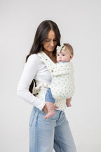 A woman with tanned skin and long, straight black hair wears a fitted white long-sleeve t-shirt and light wash wide leg denim jeans. She has a light-skinned baby with brown hair in the Mabē Monarch Buckle Carrier in Sunday Floral, which is solid white with tiny sprigs of blue flowers. The woman faces the right of the frame. The background is solid white.