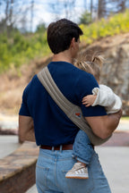 Back view of Zach Webber wearing a Mabē Monarch Toddler Sling in Charles Herringbone, which is a delicate herringbone weave cotton/linen fabric in shades of brown, cross-body with a toddler on his hip.