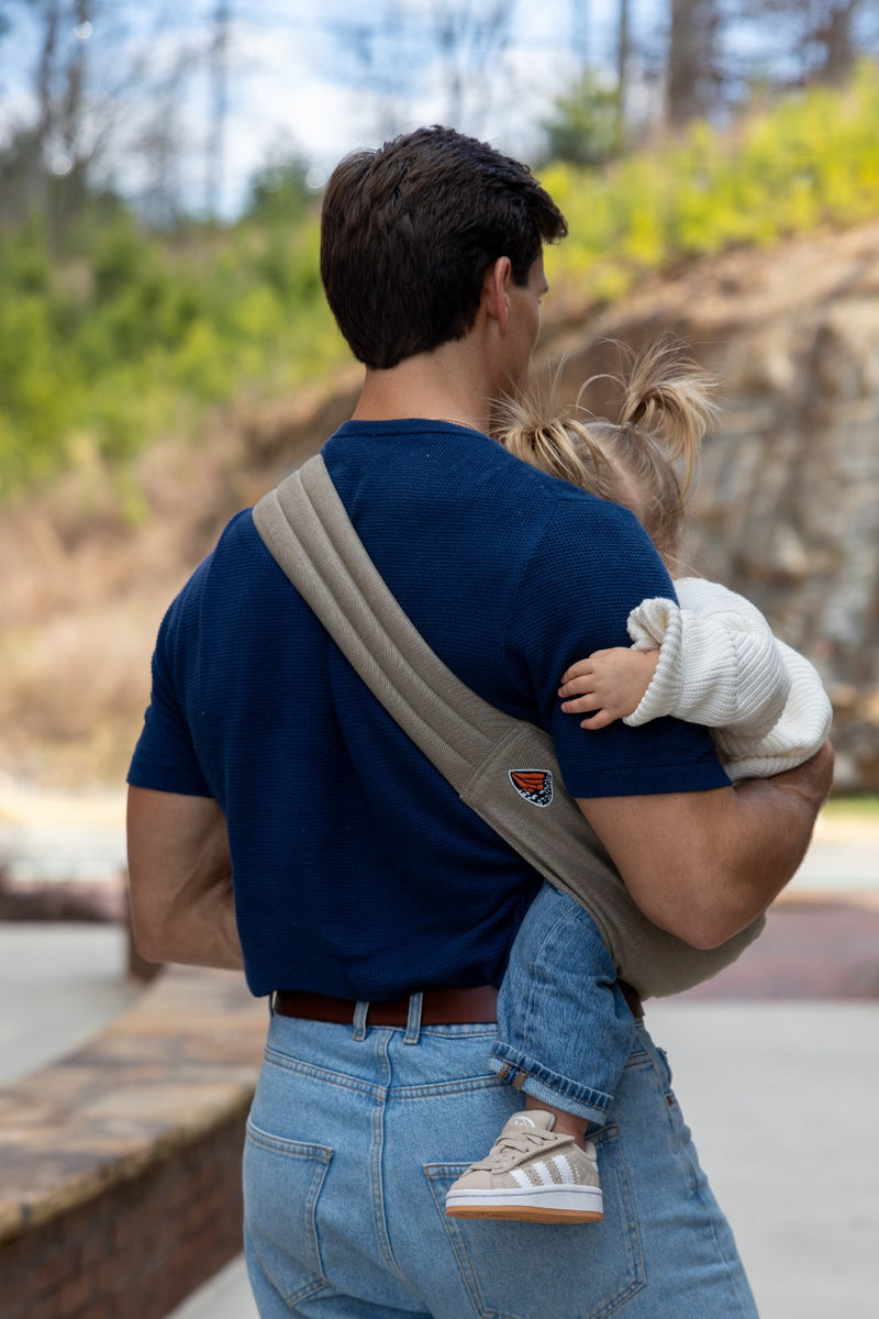 Back view of Zach Webber wearing a Mabē Monarch Toddler Sling in Charles Herringbone, which is a delicate herringbone weave cotton/linen fabric in shades of brown, cross-body with a toddler on his hip.