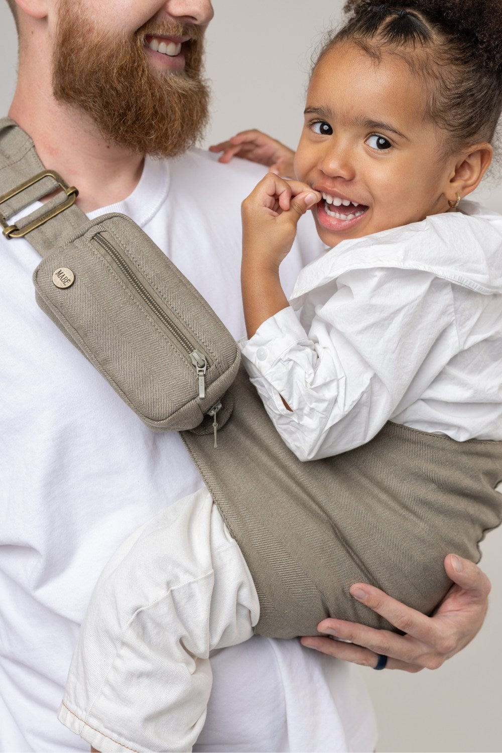 The Mabē Monarch Toddler Sling in Charles Herringbone, which is a delicate herringbone weave cotton/linen fabric in shades of brown, worn by a man with an older toddler.