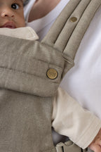 Baby in the Mabē Monarch Buckle Carrier in Charles Herringbone with an antiqued bronze button labeled 'MABĒ.' The fabric is a delicate herringbone weave in shades of brown.
