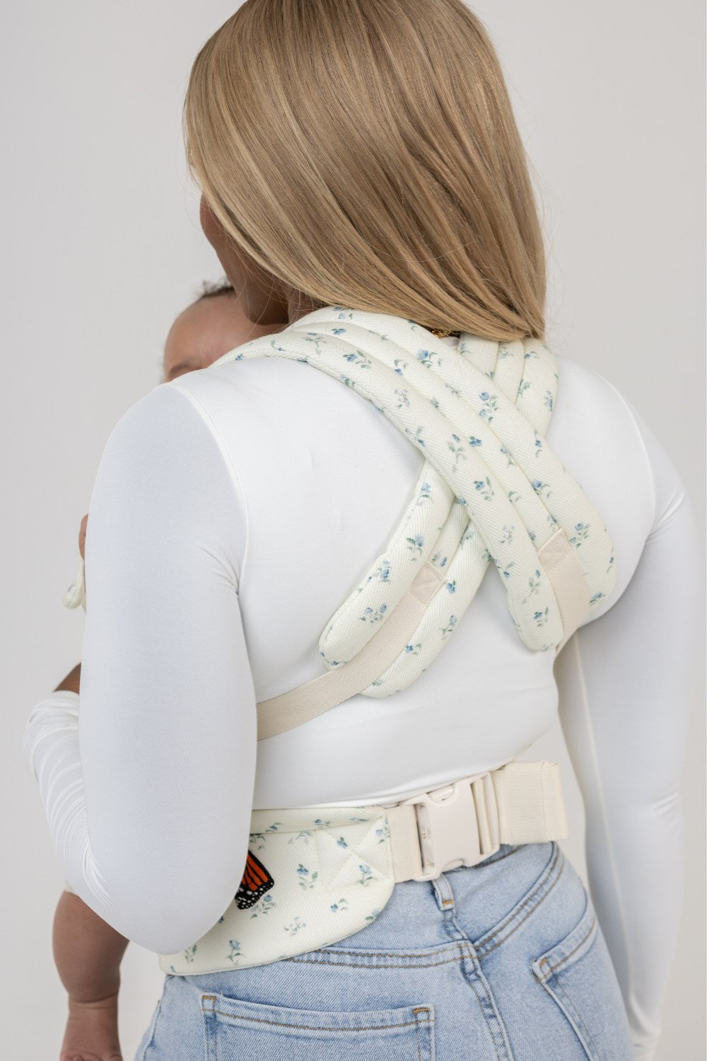 A dark-skinned woman with blonde straight hair is shown from behind wearing the Mabē Monarch Buckle Carrier in Sunday Floral, which is solid white with tiny sprigs of blue flowers. She wears a long-sleeved white shirt and light-wash denim jeans. Only the crossed straps and waistband of the carrier are visible, and one lower leg and the top of the head of a tan-skinned baby with dark hair. The background is solid white.