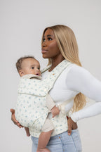 A dark-skinned woman with straight, dark-blonde hair faces the left of the frame and carries a tan-skinned baby with dark hair in the Mabē Monarch Buckle Carrier in Sunday Floral, which is solid white with tiny sprigs of blue flowers. The woman wears a fitted white long-sleeved shirt and light-wash jeans and is shown from the hips up. The background is solid white.