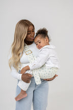 The Mabē Monarch Toddler Sling in Sunday Floral, which is solid white cotton/linen fabric with tiny sprigs of blue flowers, worn cross-body by a woman with an older toddler.