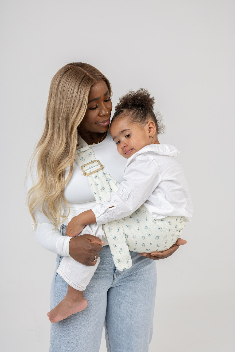 A dark-skinned woman with straight, dark-blonde hair carries a tan-skinned toddler with dark hair in the Mabē Monarch Toddler Sling in Sunday Floral, which is solid white with tiny sprigs of blue flowers. The woman wears a fitted white long-sleeved shirt and light-wash jeans and is shown from just above the knee up. The background is solid white.