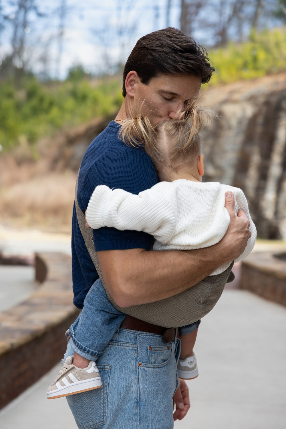 Zach Webber wearing the Mabē Monarch Toddler Sling in Charles Herringbone, which is a delicate herringbone weave cotton/linen fabric in shades of brown, with a younger toddler girl.