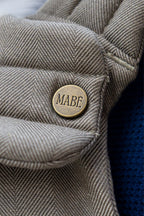 Close-up of the Mabē Monarch Buckle Carrier in Charles Herringbone with an antiqued bronze button labeled 'MABĒ.' The fabric is a delicate herringbone weave in shades of brown.