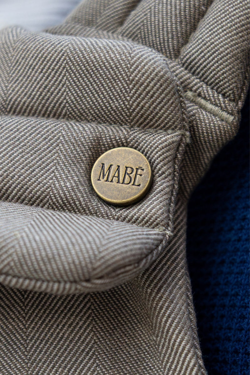 Close-up of the Mabē Monarch Buckle Carrier in Charles Herringbone with an antiqued bronze button labeled 'MABĒ.' The fabric is a delicate herringbone weave in shades of brown.