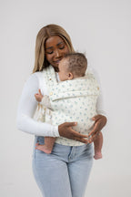 A dark-skinned woman with straight, dark-blonde hair carries a tan-skinned baby with dark hair in the Mabē Monarch Buckle Carrier in Sunday Floral, which is solid white with tiny sprigs of blue flowers. The woman wears a fitted white long-sleeved shirt and light-wash jeans and is shown from just above the knee up. She is smiling down at the baby. The background is solid white.