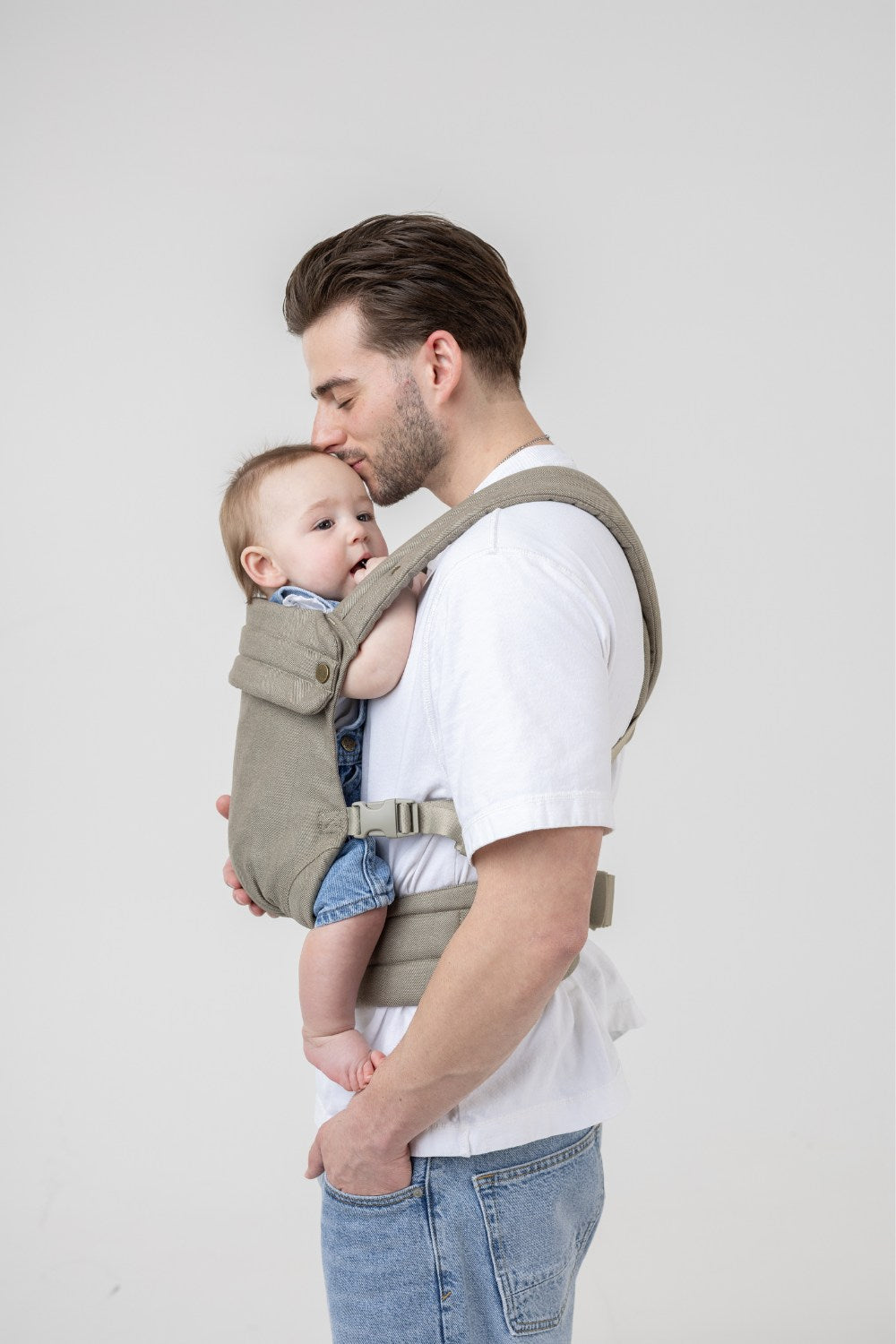 The Mabē Monarch Buckle Carrier in Charles Herringbone—a delicate herringbone weave cotton/linen fabric in shades of brown—worn by a man with a baby on his front.