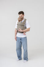 The Mabē Monarch Buckle Carrier in Charles Herringbone, which is a delicate herringbone weave cotton/linen fabric in shades of brown, worn by a man with a baby on his front.