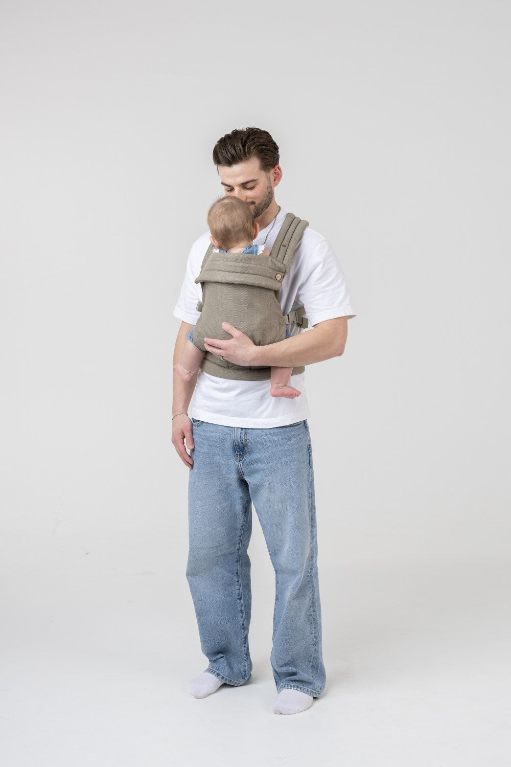 The Mabē Monarch Buckle Carrier in Charles Herringbone, which is a delicate herringbone weave cotton/linen fabric in shades of brown, worn by a man with a baby on his front.