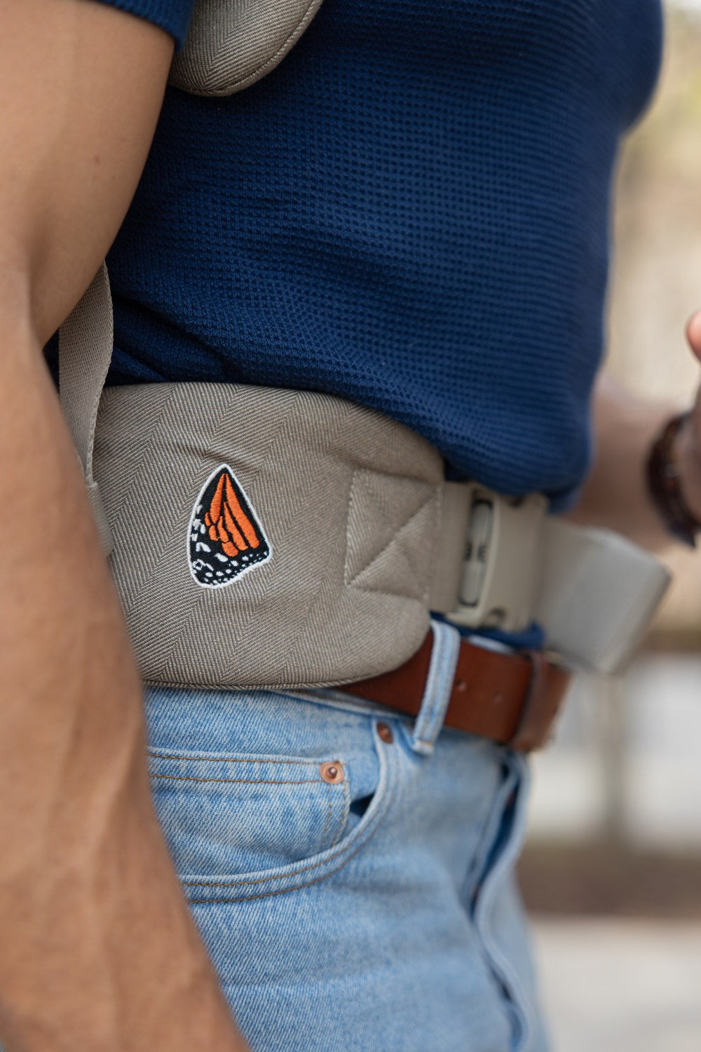 A detail photo of an unseen person wearing a Mabē Monarch Buckle Carrier in Charles Herringbone, which is a delicate herringbone weave in shades of brown. Only the front of the waistband of the carrier, which is being worn in backpack mode, is visible, with some navy blue shirt above and some light-wash jeans below. A tanned arm is visible in the foreground and the background is blurry.