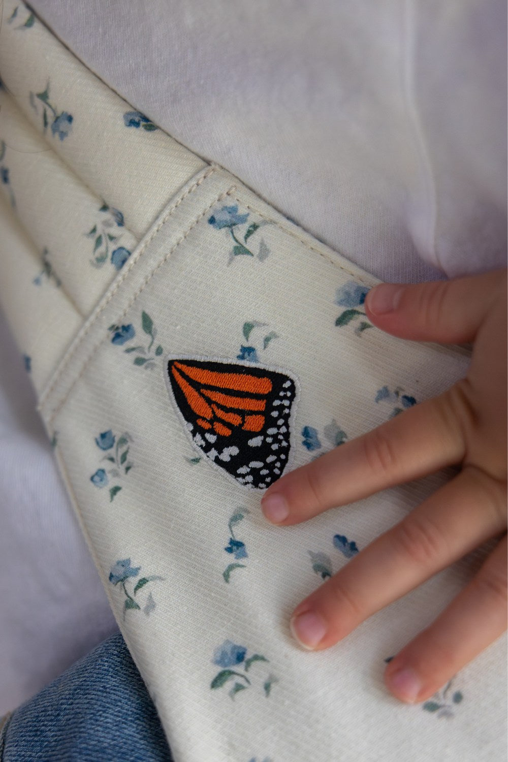 A child's hand over the top of a detail shot of the Mabē Monarch Toddler Sling in Sunday Floral, which is solid white cotton/linen fabric with tiny springs of blue flowers and an embroidered black and orange butterfly wing.