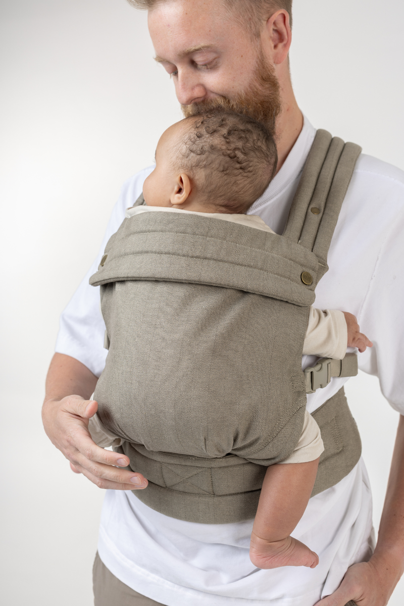 Man wearing the Mabē Monarch Buckle Carrier in Charles Herringbone—a delicate herringbone weave in shades of brown—to carry a baby.