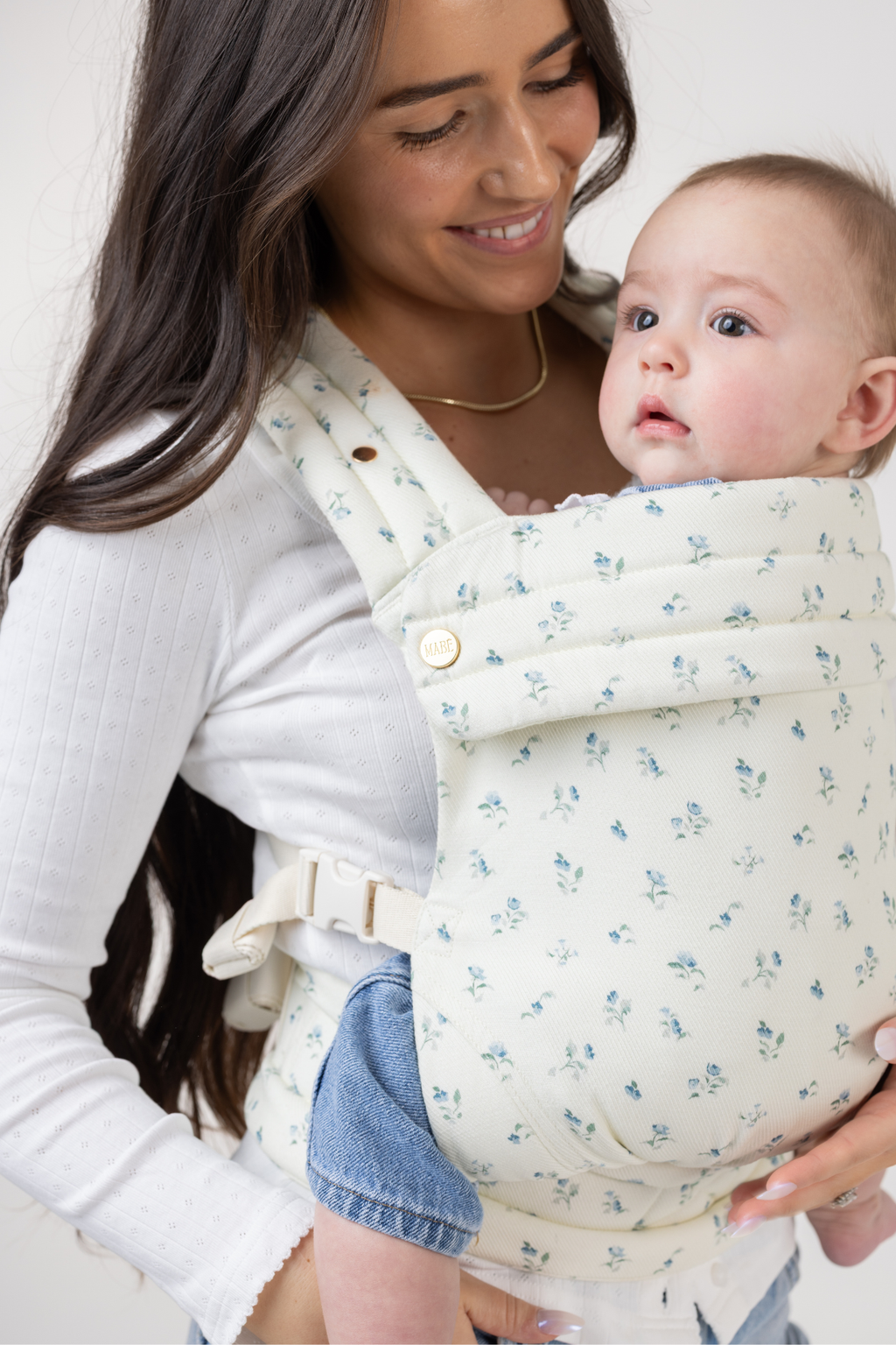 Angled view of a tan-skinned woman with very long dark brown hair with slight waves. She wears a long-sleeved white shirt and light-wash denim jeans. She is shown from the hips up and wears a light-skinned baby with light brown hair and a headband in the Mabē Monarch Buckle Carrier in Sunday Floral, which is white with tiny sprigs of blue flowers. The baby faces left of the frame.