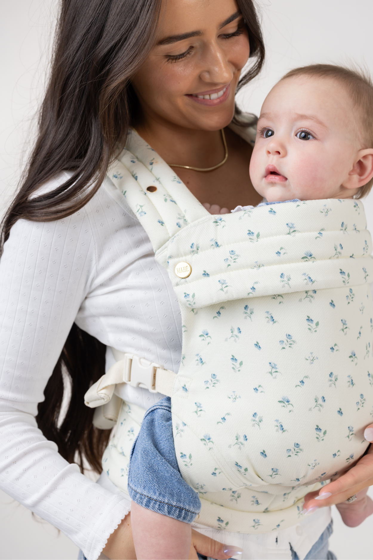 Angled view of a tan-skinned woman with very long dark brown hair with slight waves. She wears a long-sleeved white shirt and light-wash denim jeans. She is shown from the hips up and wears a light-skinned baby with light brown hair and a headband in the Mabē Monarch Buckle Carrier in Sunday Floral, which is white with tiny sprigs of blue flowers. The baby faces left of the frame.