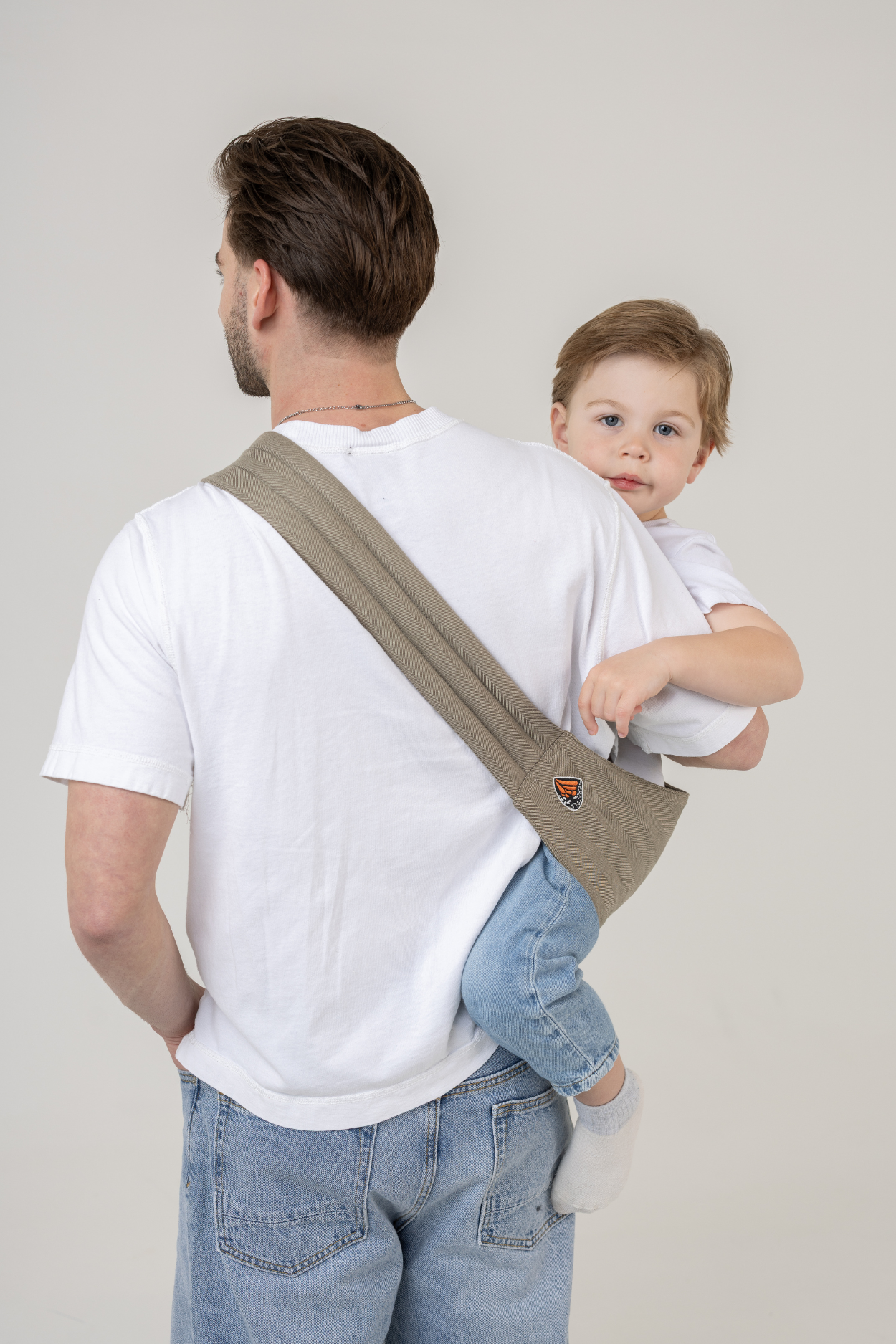 Man wearing the Mabē Monarch Toddler Sling in Charles Herringbone—delicate herringbone weave cotton/linen fabric in shades of brown—to carry a toddler boy.