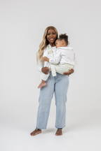 Woman wearing the Mabē Monarch Toddler Sling in Sunday Floral—solid white cotton/linen fabric with tiny sprigs of blue flowers and a gold buckle—to carry a toddler girl.