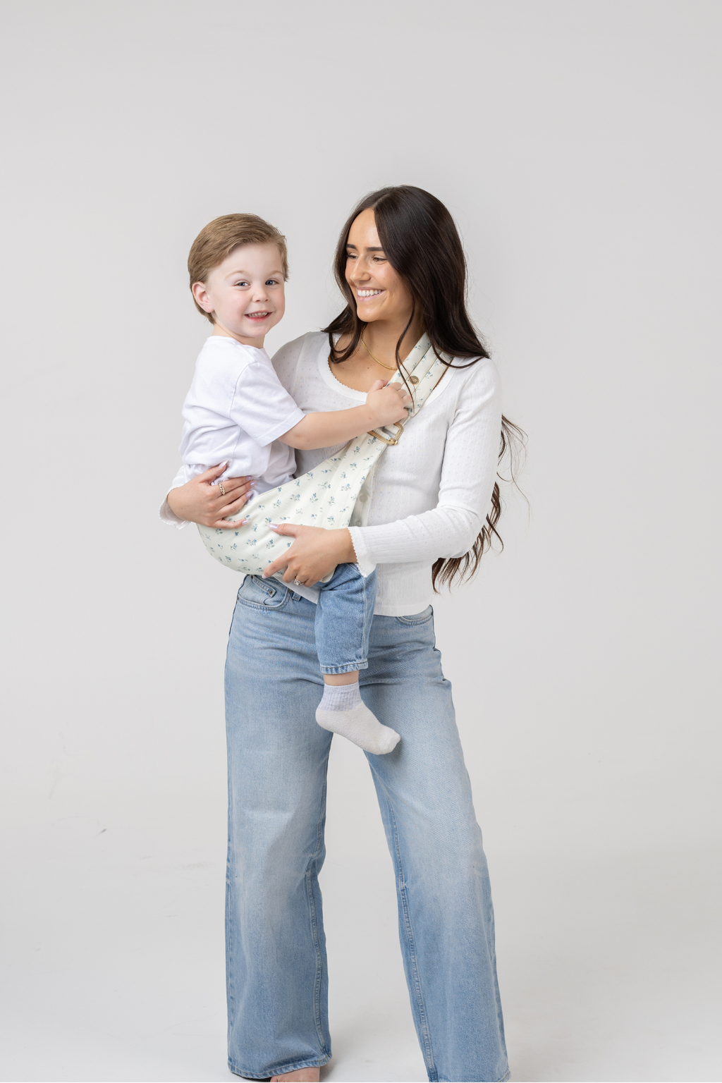 Woman wearing the Mabē Monarch Buckle Carrier in Sunday Floral—white cotton/linen fabric with tiny sprigs of blue flowers—to carry a toddler boy.