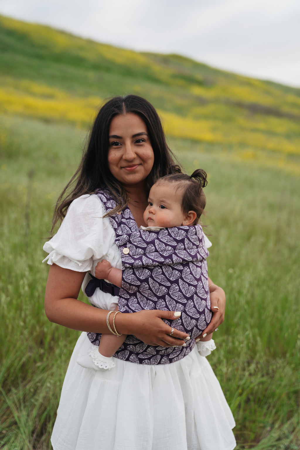 A mom stands in a green field wearing her baby in the Mabē Monarch Baby Carrier in Violet Wings, a hand-drawn deep purple butterfly print.