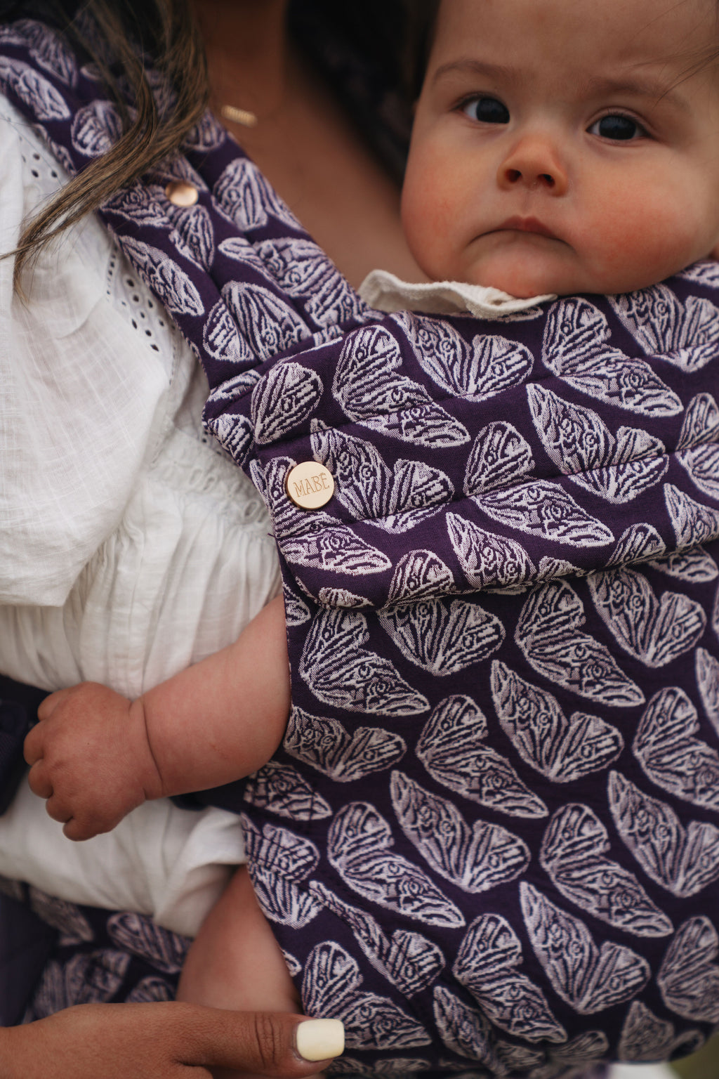 Close-up of the hand-drawn butterfly print on the Mabē Monarch Baby Carrier in Violet Wings, with a baby peeking out.