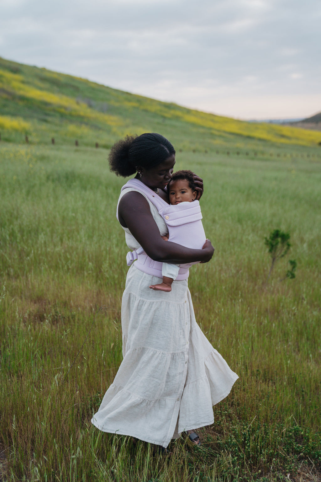 A mom in a long white dress stands in a green field wearing her baby in the Mabē Monarch Baby Carrier in Lilac Skies.