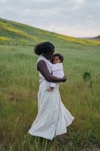 A mom in a long white dress stands in a green field wearing her baby in the Mabē Monarch Baby Carrier in Lilac Skies.
