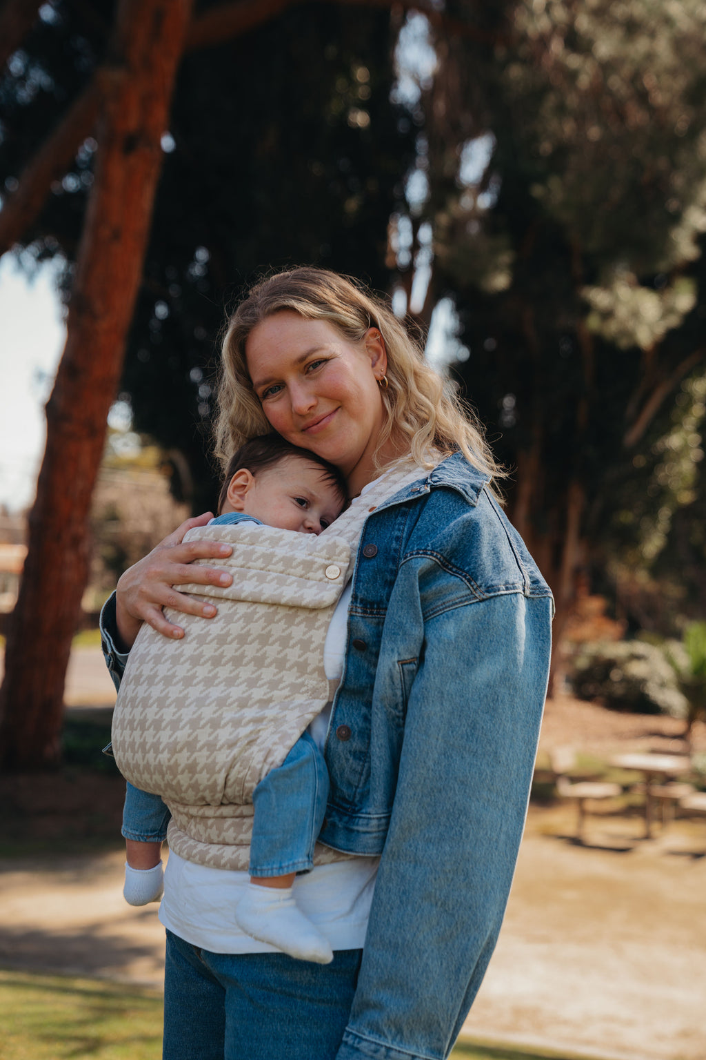 A mom in a denim jacket wears her baby in the Mabē Monarch Baby Carrier in Houndstooth, front carry, outside under the trees.