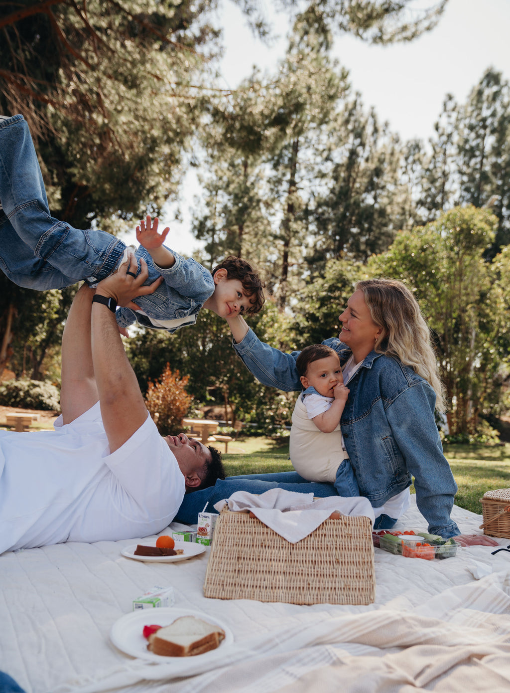 A family picnic on a blanket outdoors, with mom wearing her baby in the Mabē Monarch Baby Carrier in Natural Ivory while dad lifts an older child.