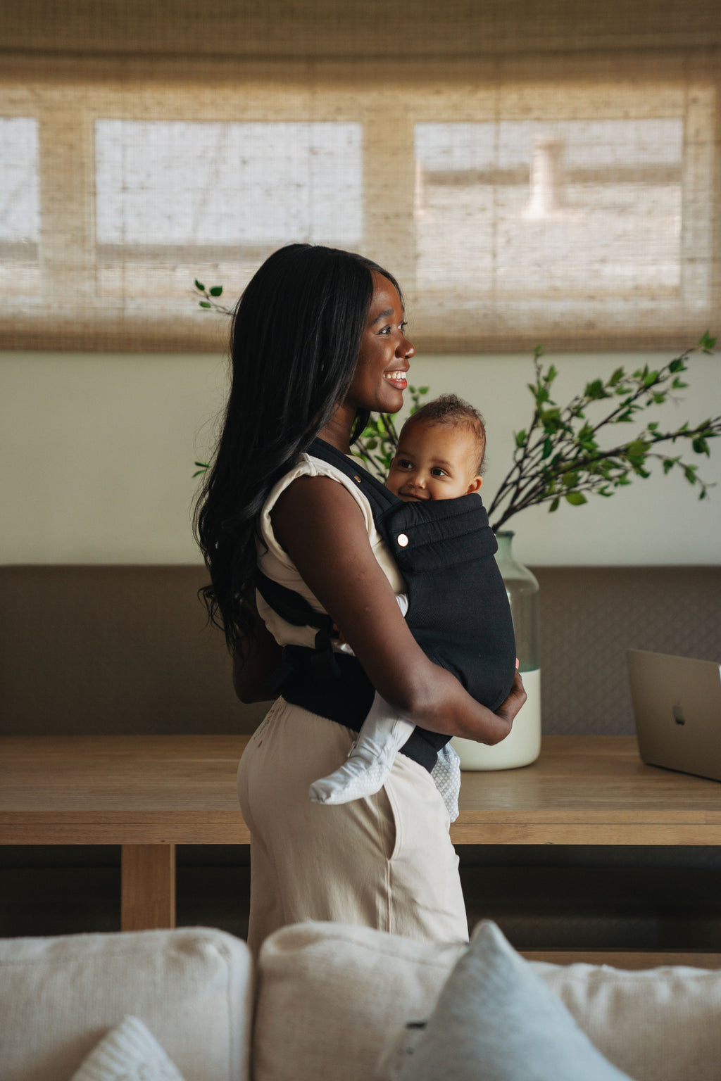 A mom stands in a bright living room wearing her baby in the Mabē Monarch Baby Carrier in Ebony, side view.