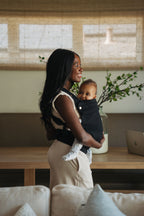 A mom stands in a bright living room wearing her baby in the Mabē Monarch Baby Carrier in Ebony, side view.