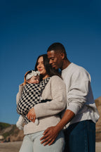 A couple embraces outdoors under a blue sky while mom wears the baby in the Mabē Monarch Baby Carrier in Ebony Check.