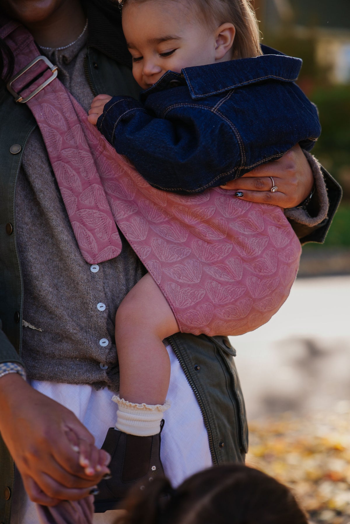 A toddler rests on mom's hip in the Mabē Monarch Toddler Sling in Mauve Wings, close-up of the blush pink butterfly print.