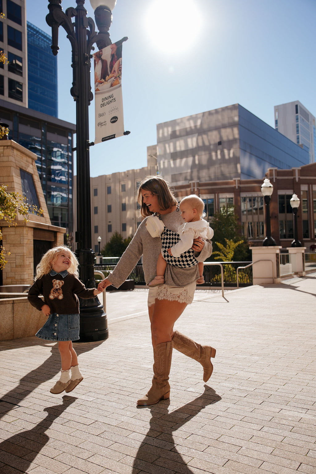A mom walks through a sunny city plaza with her toddler in the Mabē Monarch Toddler Sling in Ebony Check while her older child walks beside her.