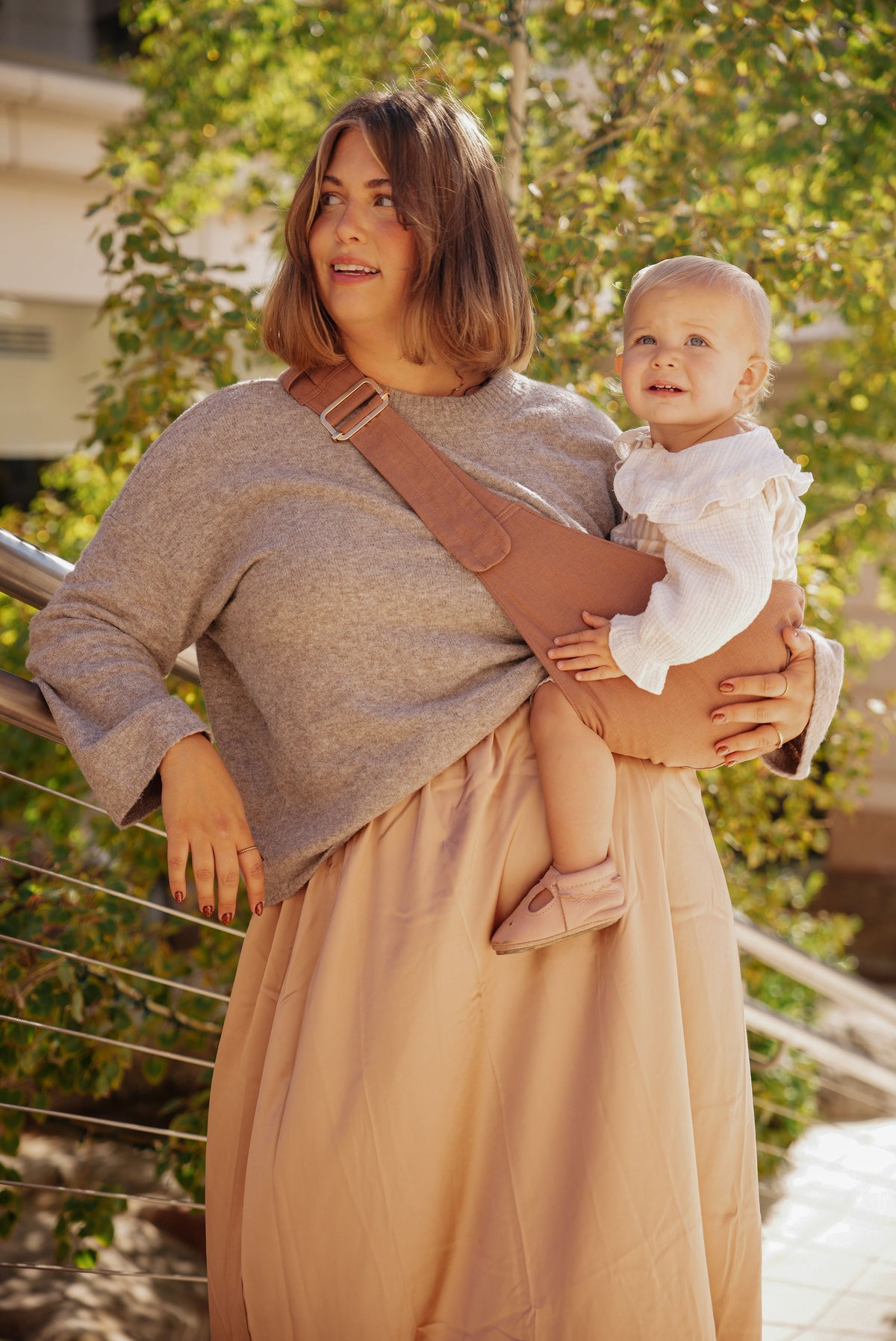 A mom outdoors carries her smiling baby on her hip in the Mabē Monarch Toddler Sling in Mocha.