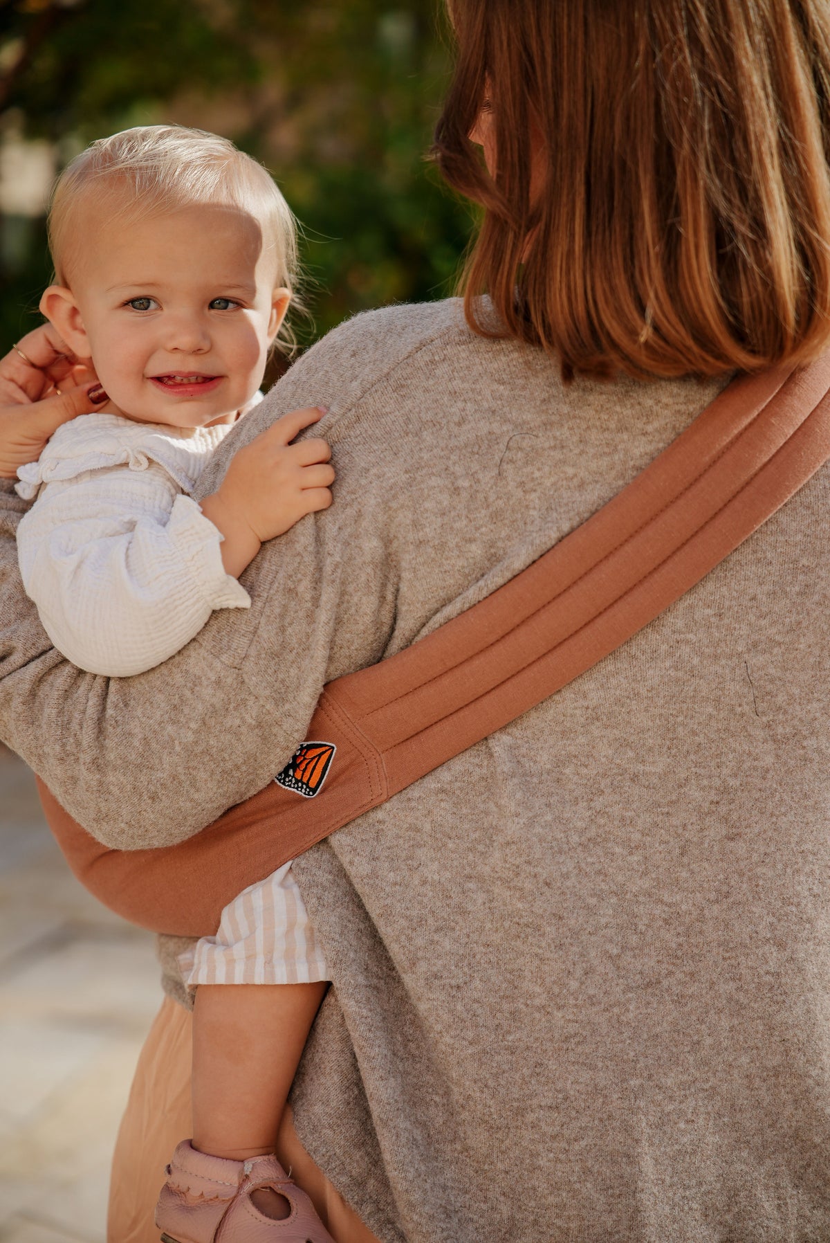 Back view of a mom carrying her toddler in the Mabē Monarch Toddler Sling in Mocha, showing the strap across her back.