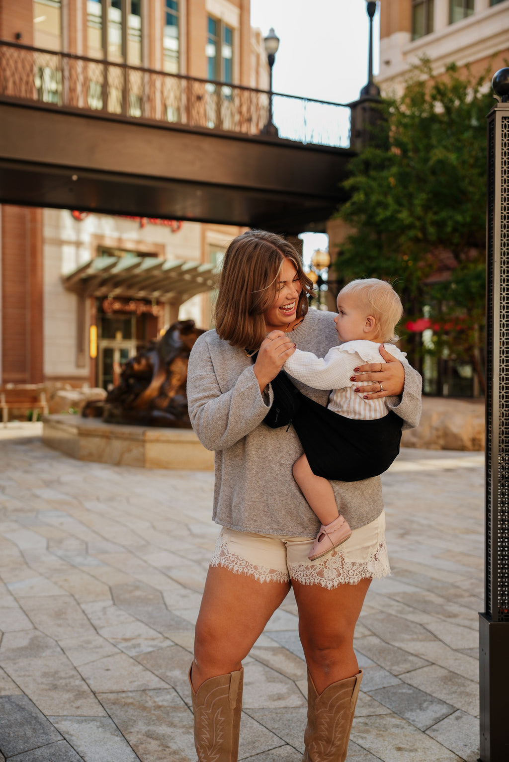 A mom walks through a city plaza carrying her toddler in the Mabē Monarch Toddler Sling in Ebony, full length.