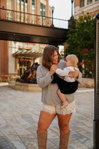 A mom walks through a city plaza carrying her toddler in the Mabē Monarch Toddler Sling in Ebony, full length.