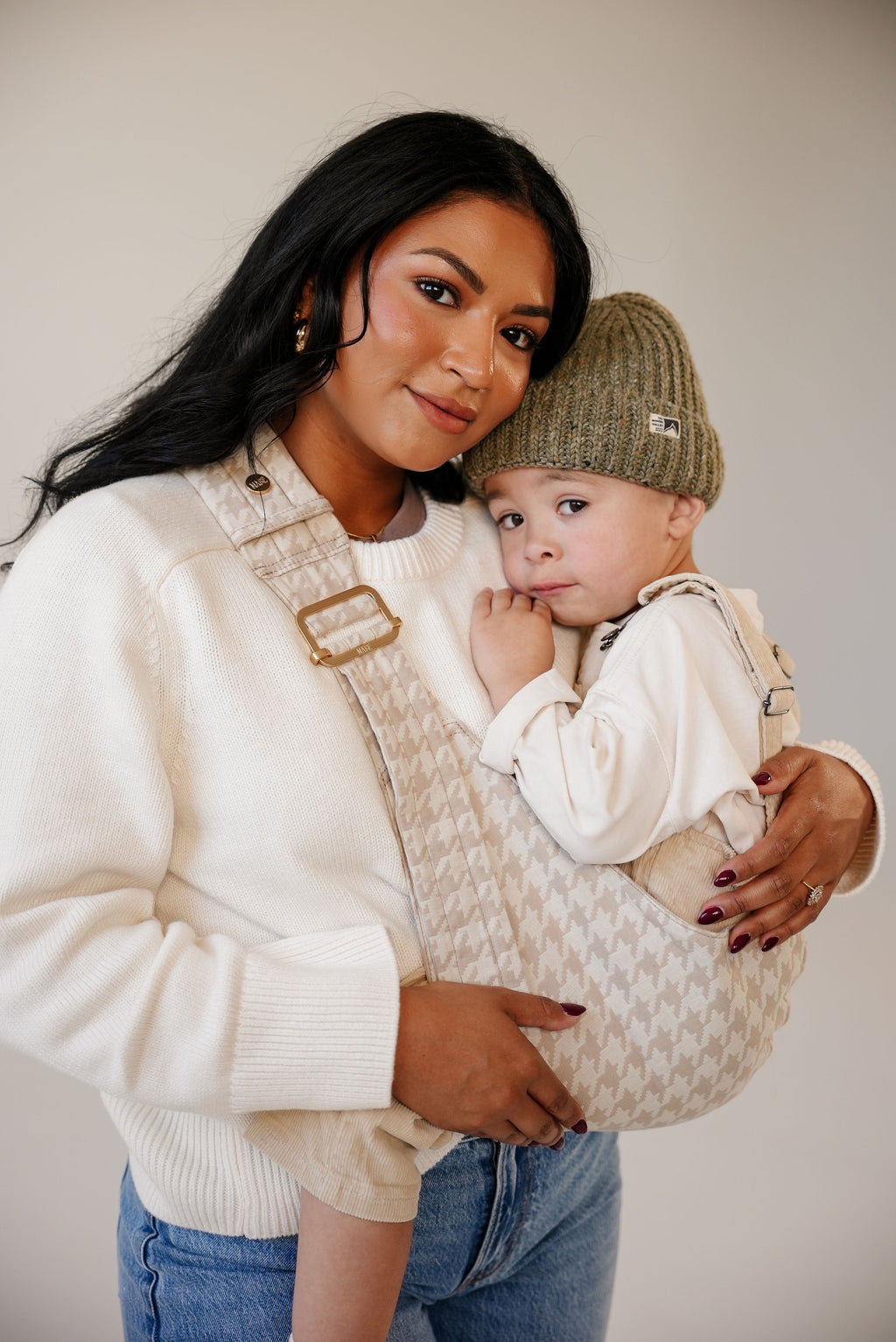A mom holds her toddler on her hip in the Mabē Monarch Toddler Sling in Houndstooth against a neutral studio backdrop.