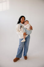 A mom holds her toddler in the Mabē Monarch Toddler Sling in Dream Wash Denim against a white studio background, full length.