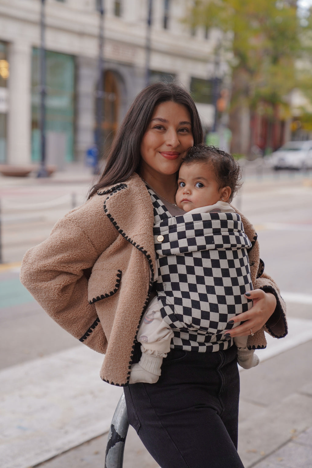 A mom wears her baby in the Mabē Monarch Baby Carrier in Ebony Check on a city street.